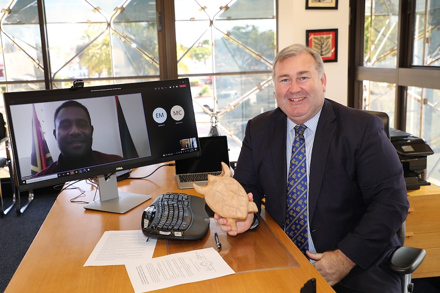 A white middle aged man smiles at the camera sitting next to a computer screen where he's skyping with a Vanuatu man   
